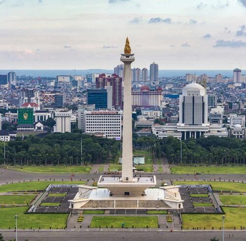 National Monument (Monas) in Jakarta, Indonesia, standing tall among the modern city skyline with skyscrapers and the golden flame monument visible during sunset.
