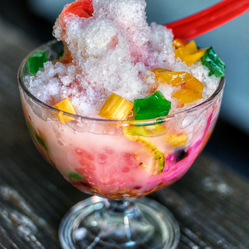 Colorful Indonesian es campur dessert served in a glass bowl with shaved ice, jelly cubes, fruit slices, and sweet milk syrup on a dark wooden table.