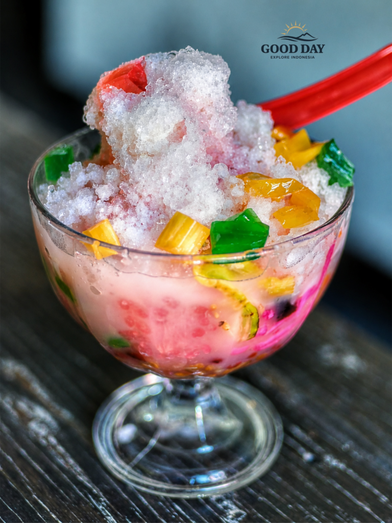 Colorful Indonesian es campur dessert served in a glass bowl with shaved ice, jelly cubes, fruit slices, and sweet milk syrup on a dark wooden table.