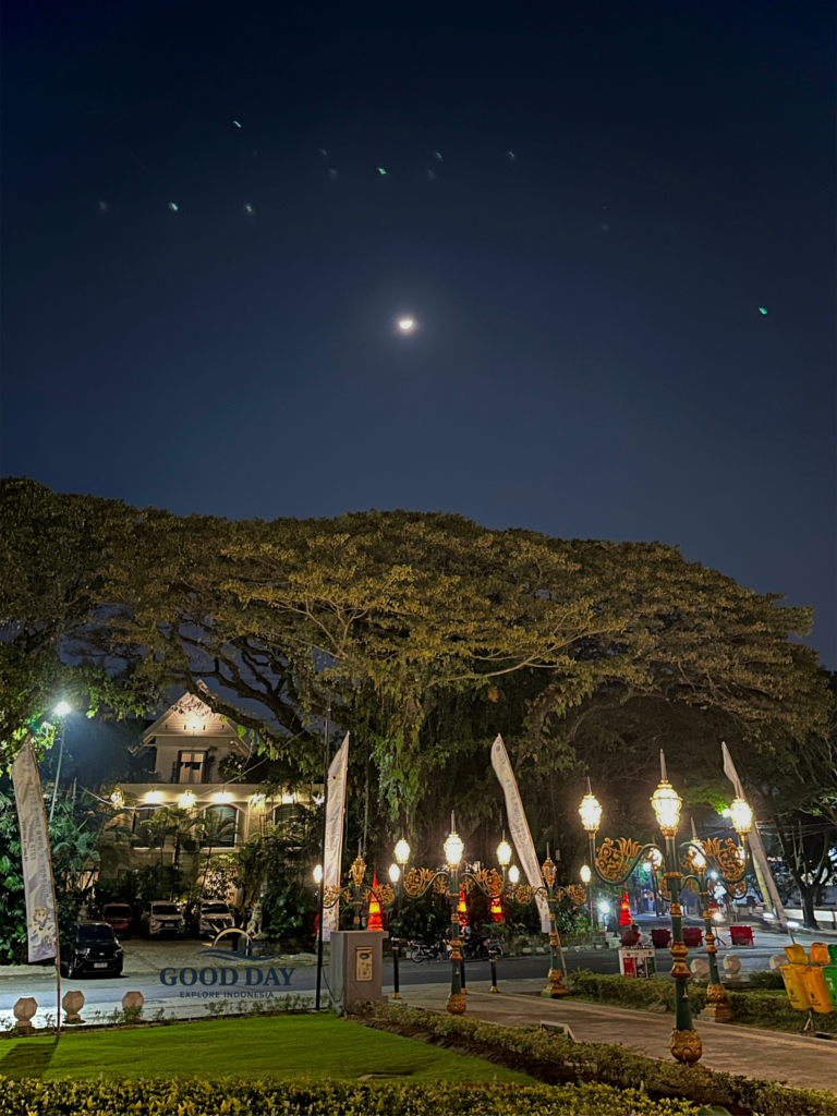 Beautiful night view of Balai Kota Malang with glowing vintage street lamps, giant shady trees, and a bright moon illuminating the peaceful city atmosphere in Malang, Indonesia.
