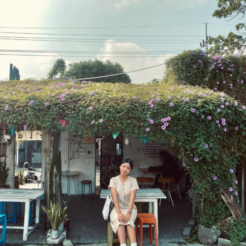Young woman sitting in front of a cozy rustic café covered with blooming purple flowers under a bright blue sky, creating a peaceful aesthetic street café atmosphere in Indonesia.