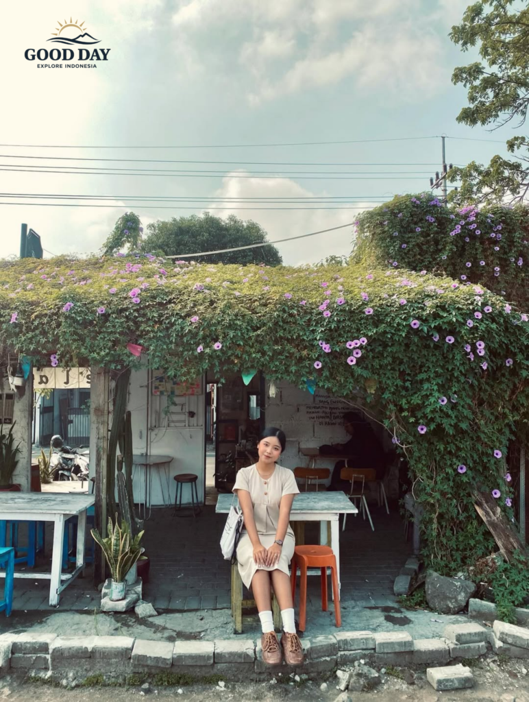 Young woman sitting in front of a cozy rustic café covered with blooming purple flowers under a bright blue sky, creating a peaceful aesthetic street café atmosphere in Indonesia.