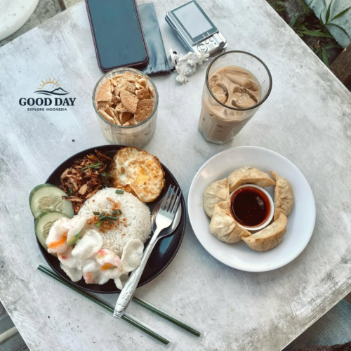 Top-view aesthetic Indonesian breakfast setup on a rustic white table featuring nasi putih with fried egg, shredded chicken, crackers, cucumber slices, dumplings with chili sauce, iced coffee, and biscuit dessert alongside a smartphone and vintage camera in an outdoor café setting.