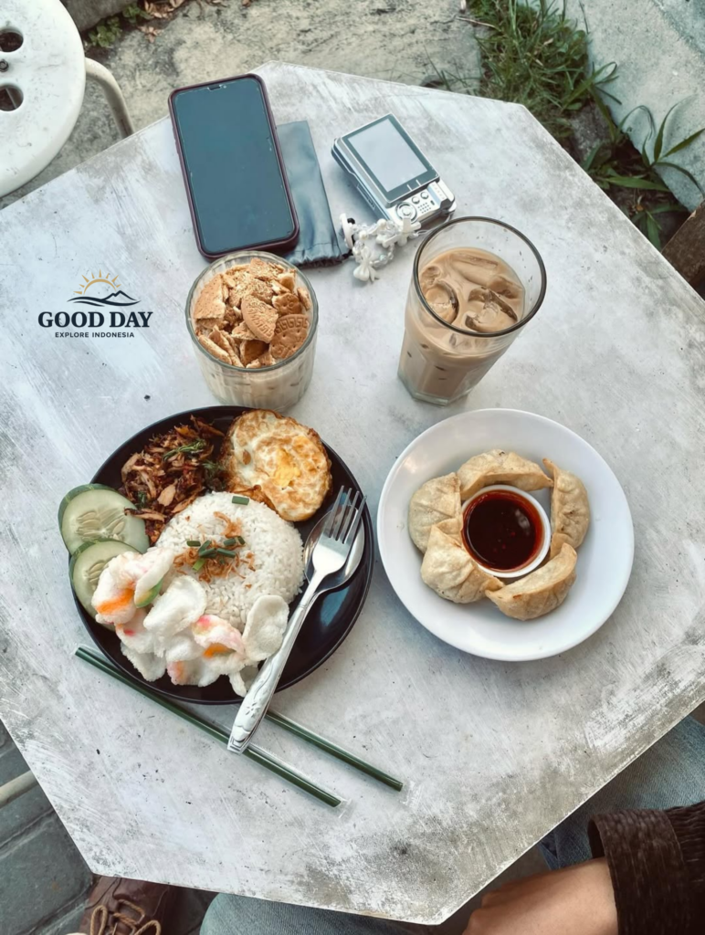 Top-view aesthetic Indonesian breakfast setup on a rustic white table featuring nasi putih with fried egg, shredded chicken, crackers, cucumber slices, dumplings with chili sauce, iced coffee, and biscuit dessert alongside a smartphone and vintage camera in an outdoor café setting.