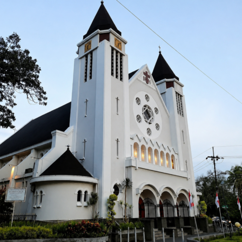 Beautiful white Catholic church with twin towers and black rooftops viewed from the street during sunset in Indonesia, featuring classic European-inspired architecture and peaceful evening atmosphere.