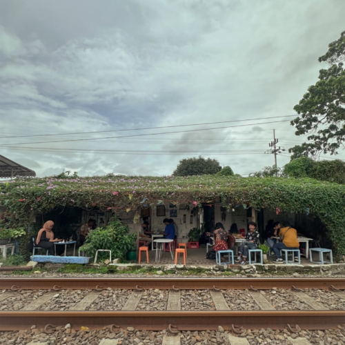 elak Kopi Malang, a unique railway-side café in Malang, Indonesia, where visitors relax and enjoy coffee beside active train tracks. Surrounded by lush greenery and a cozy open-air atmosphere, the café offers a distinctive local experience under dramatic cloudy skies.