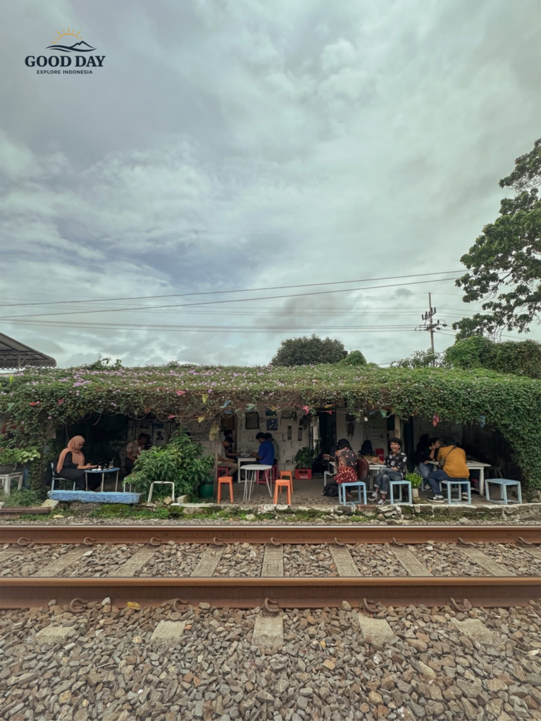 elak Kopi Malang, a unique railway-side café in Malang, Indonesia, where visitors relax and enjoy coffee beside active train tracks. Surrounded by lush greenery and a cozy open-air atmosphere, the café offers a distinctive local experience under dramatic cloudy skies.