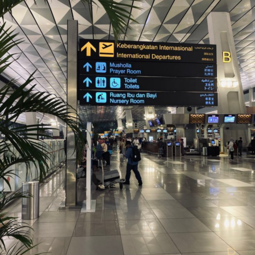 Interior of Soekarno-Hatta International Airport in Jakarta showing international departures signage, modern terminal design, travelers with luggage, and airport facilities including prayer room and toilets.