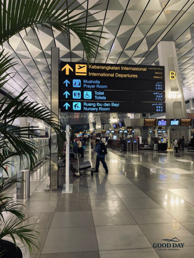 Interior of Soekarno-Hatta International Airport in Jakarta showing international departures signage, modern terminal design, travelers with luggage, and airport facilities including prayer room and toilets.