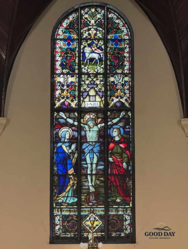 Stained glass window inside the Jakarta Catholic Cathedral featuring the crucifixion of Jesus Christ, with intricate Gothic-style religious artwork, vibrant blue and red colors, and sacred Christian symbolism in Jakarta, Indonesia