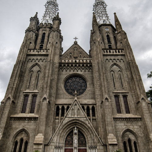 Front exterior view of Jakarta Catholic Cathedral featuring Neo-Gothic architecture, tall twin spires, arched entrances, and historic stone facade under a cloudy sky in Jakarta, Indonesia