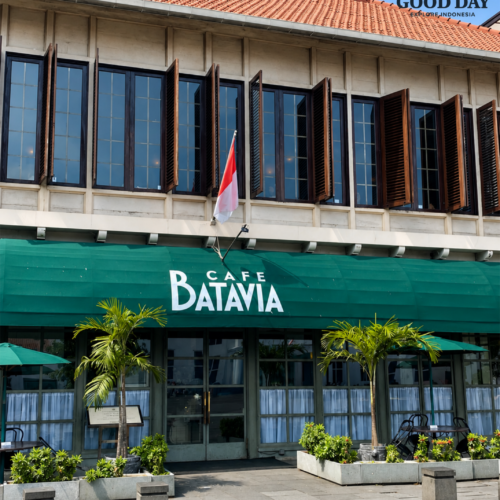 Exterior view of Café Batavia in Jakarta Old Town (Kota Tua), Indonesia, featuring colonial Dutch architecture, large wooden shutter windows, a green café awning, tropical plants, and the Indonesian flag under a bright blue sky.