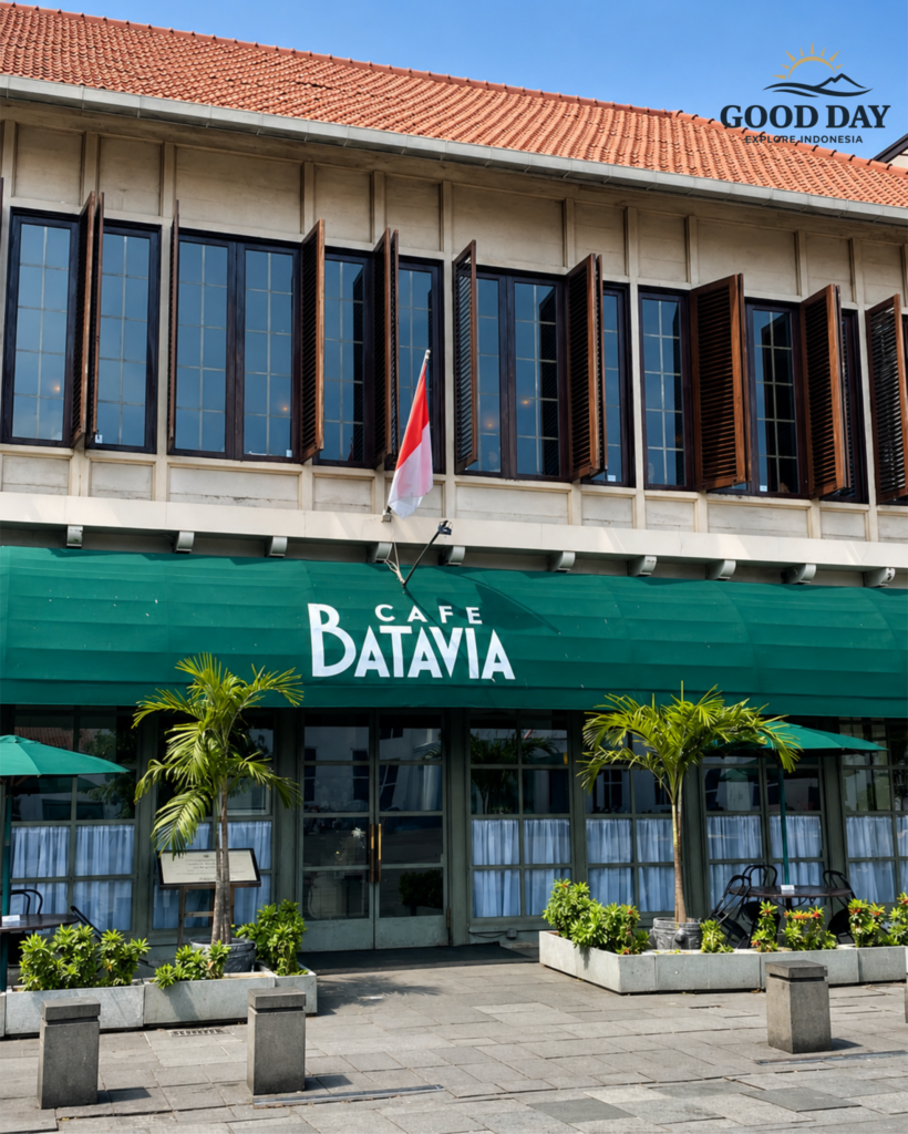 Exterior view of Café Batavia in Jakarta Old Town (Kota Tua), Indonesia, featuring colonial Dutch architecture, large wooden shutter windows, a green café awning, tropical plants, and the Indonesian flag under a bright blue sky.