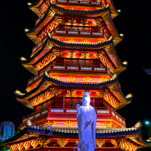 Colorfully illuminated Chinese-style pagoda at PIK 2 Jakarta at night, featuring traditional architecture, vibrant red and gold lighting, a large Buddhist statue, and visitors exploring the cultural attraction.
