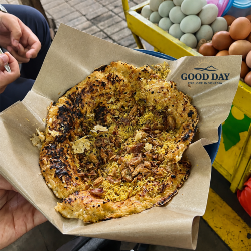 Traditional Jakarta Kerak Telor freshly cooked over charcoal, topped with crispy shredded coconut and fried shallots, served on food-grade paper beside a street food cart.