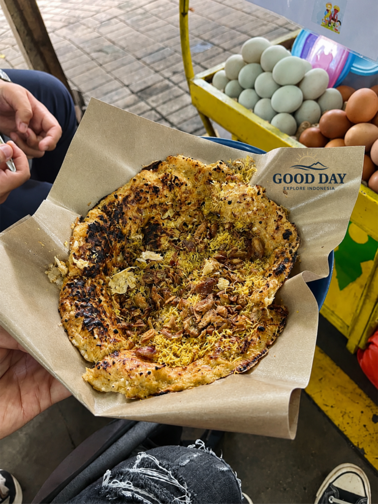 Traditional Jakarta Kerak Telor freshly cooked over charcoal, topped with crispy shredded coconut and fried shallots, served on food-grade paper beside a street food cart.