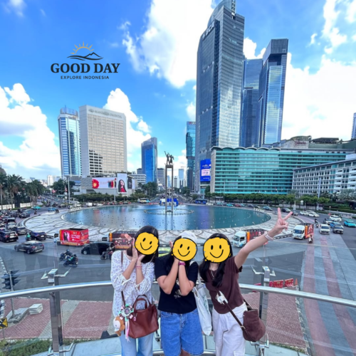 Three visitors posing at Bundaran HI Jakarta with the iconic Selamat Datang Monument and modern skyscrapers in the background. The vibrant cityscape showcases Jakarta's bustling business district, famous roundabout, and contemporary urban skyline under a bright blue sky.