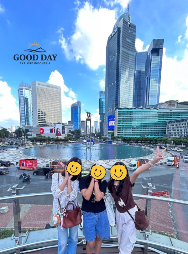 Three visitors posing at Bundaran HI Jakarta with the iconic Selamat Datang Monument and modern skyscrapers in the background. The vibrant cityscape showcases Jakarta's bustling business district, famous roundabout, and contemporary urban skyline under a bright blue sky.