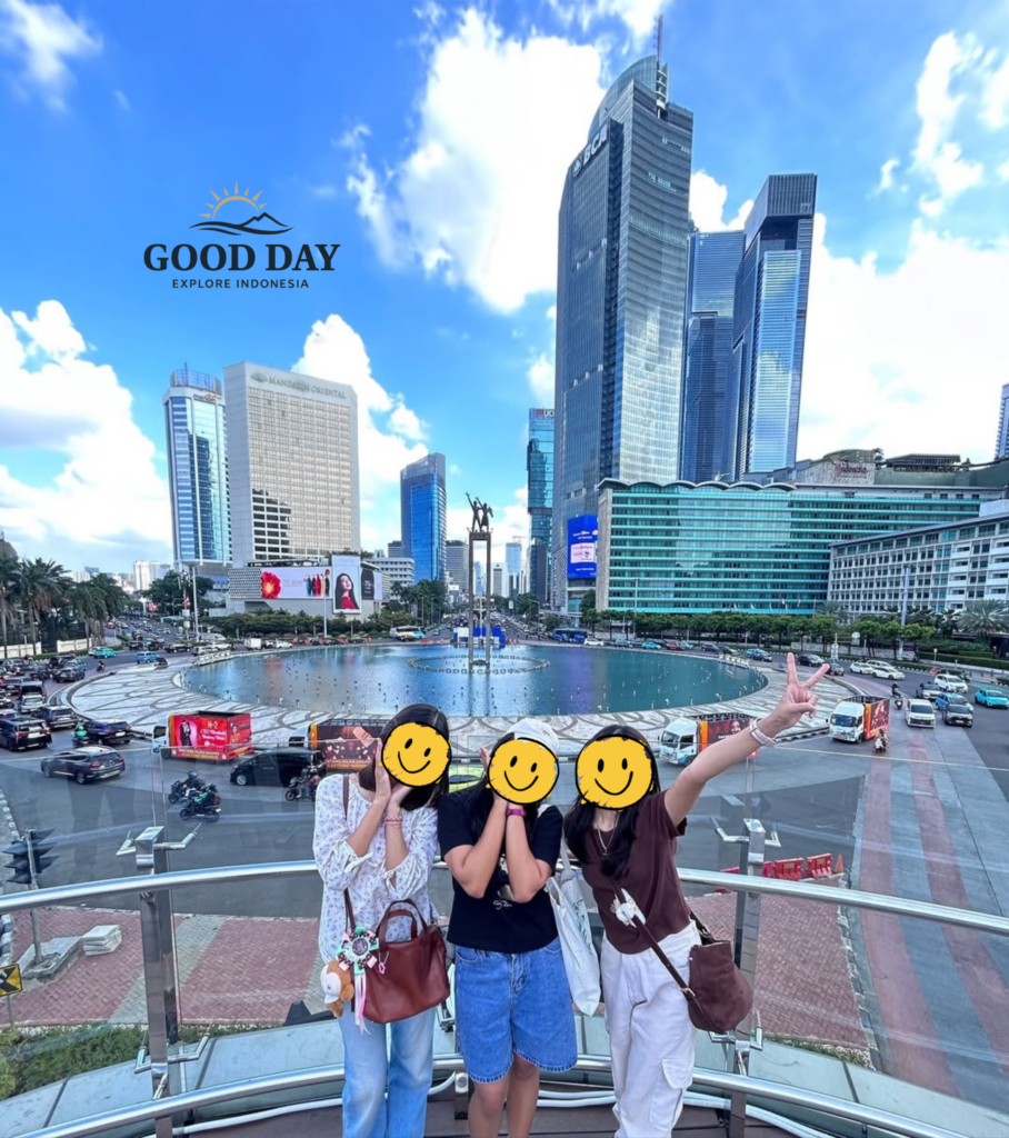 Three visitors posing at Bundaran HI Jakarta with the iconic Selamat Datang Monument and modern skyscrapers in the background. The vibrant cityscape showcases Jakarta's bustling business district, famous roundabout, and contemporary urban skyline under a bright blue sky.