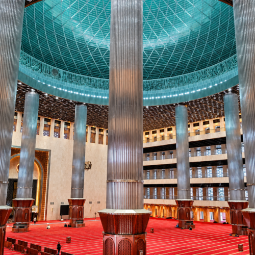 Magnificent prayer hall inside Istiqlal Mosque Jakarta featuring towering stainless-steel columns, a massive turquoise dome, intricate Islamic calligraphy, and expansive red prayer carpets. The grand interior showcases the architectural beauty of Indonesia's national mosque and the largest mosque in Southeast Asia.