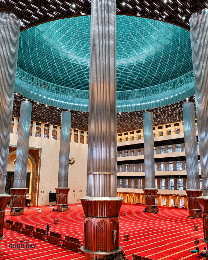 Magnificent prayer hall inside Istiqlal Mosque Jakarta featuring towering stainless-steel columns, a massive turquoise dome, intricate Islamic calligraphy, and expansive red prayer carpets. The grand interior showcases the architectural beauty of Indonesia's national mosque and the largest mosque in Southeast Asia.