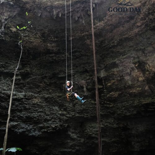 Adventurer descending by rope inside Goa Macan Mati, Yogyakarta, Indonesia. Suspended in the middle of a massive vertical cave chamber, the participant uses professional caving equipment while surrounded by towering limestone walls, hanging roots, and the dramatic underground landscape of Gunungkidul