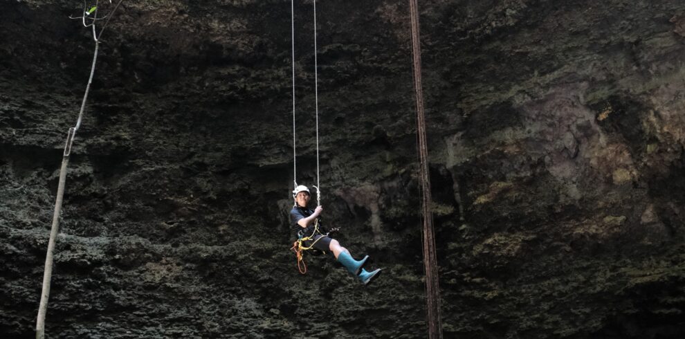 Adventurer descending by rope inside Goa Macan Mati, Yogyakarta, Indonesia. Suspended in the middle of a massive vertical cave chamber, the participant uses professional caving equipment while surrounded by towering limestone walls, hanging roots, and the dramatic underground landscape of Gunungkidul