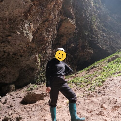 Visitor standing inside Goa Macan Mati in Yogyakarta, Indonesia, surrounded by towering limestone walls and illuminated by natural sunlight entering through the cave opening above. Wearing safety gear and boots suitable for cave exploration, the adventurer experiences the breathtaking scale of one of Gunungkidul's most impressive underground destinations.