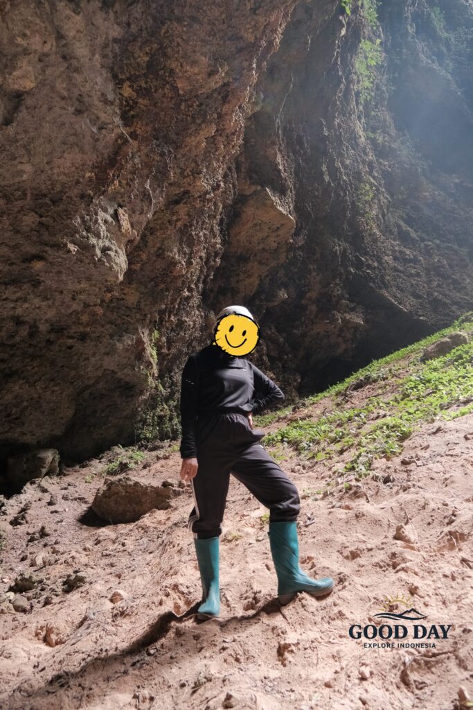 Visitor standing inside Goa Macan Mati in Yogyakarta, Indonesia, surrounded by towering limestone walls and illuminated by natural sunlight entering through the cave opening above. Wearing safety gear and boots suitable for cave exploration, the adventurer experiences the breathtaking scale of one of Gunungkidul's most impressive underground destinations.