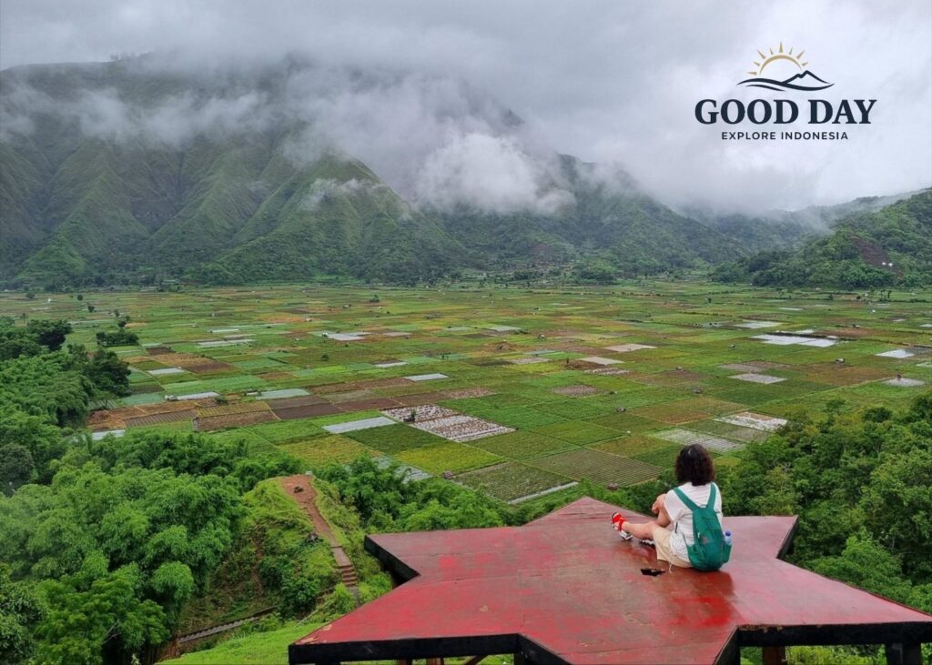 Traveler sitting on a star-shaped viewpoint overlooking the Sembalun Valley in Lombok, Indonesia, surrounded by patchwork farmland, misty green mountains, and dramatic low clouds.