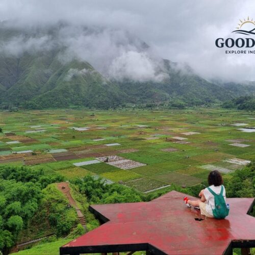 Traveler sitting on a star-shaped viewpoint overlooking the Sembalun Valley in Lombok, Indonesia, surrounded by patchwork farmland, misty green mountains, and dramatic low clouds.