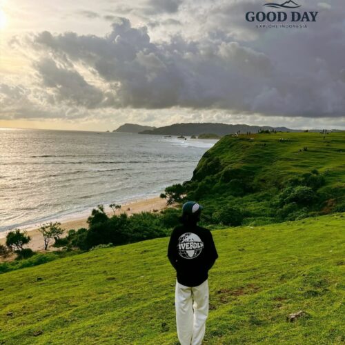 Visitor standing on the lush green hills of Merese Hill in Lombok, Indonesia, overlooking dramatic coastal cliffs, pristine beaches, and the vast Indian Ocean under a dramatic cloudy sky. The viewpoint offers one of the most breathtaking sunset panoramas in the Mandalika region.