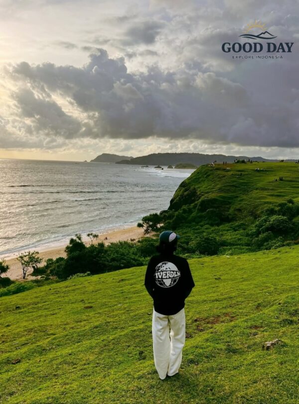 Visitor standing on the lush green hills of Merese Hill in Lombok, Indonesia, overlooking dramatic coastal cliffs, pristine beaches, and the vast Indian Ocean under a dramatic cloudy sky. The viewpoint offers one of the most breathtaking sunset panoramas in the Mandalika region.
