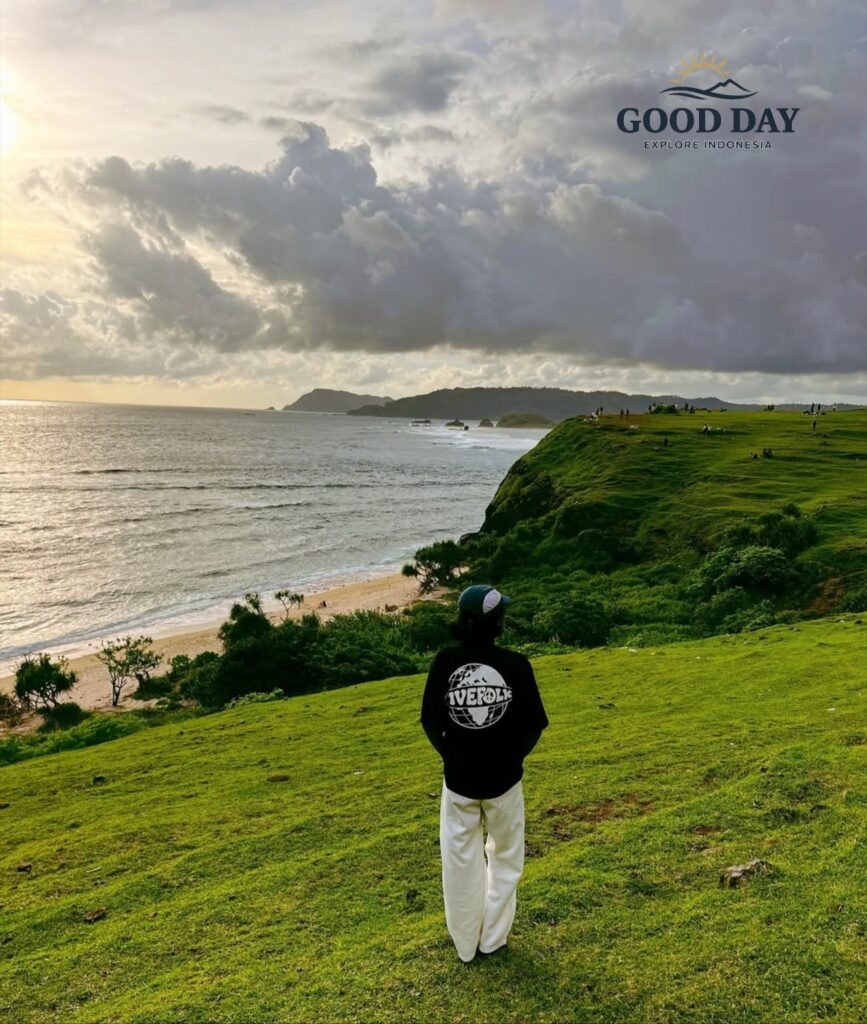 Visitor standing on the lush green hills of Merese Hill in Lombok, Indonesia, overlooking dramatic coastal cliffs, pristine beaches, and the vast Indian Ocean under a dramatic cloudy sky. The viewpoint offers one of the most breathtaking sunset panoramas in the Mandalika region.