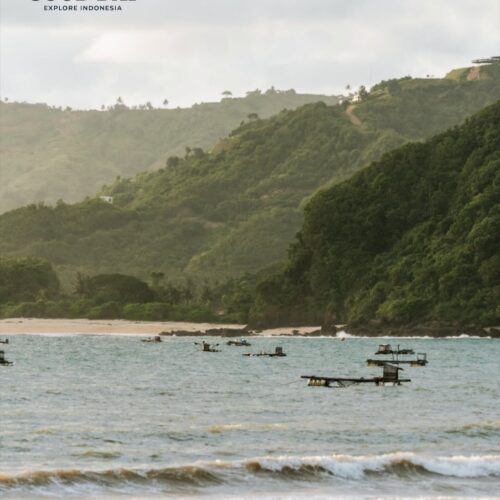 Lombok beach with gentle waves, traditional fishing platforms floating offshore, and lush green hills rising along the coastline under a soft tropical sky in West Nusa Tenggara, Indonesia.