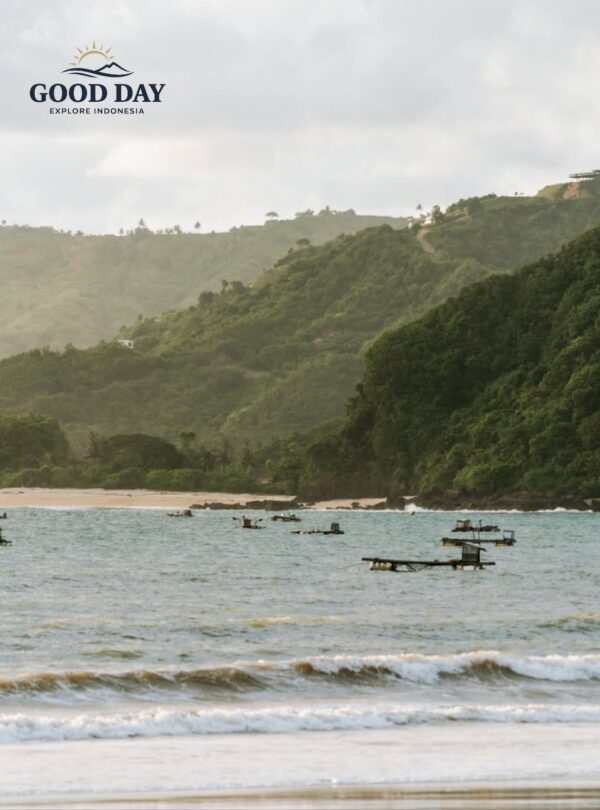 Lombok beach with gentle waves, traditional fishing platforms floating offshore, and lush green hills rising along the coastline under a soft tropical sky in West Nusa Tenggara, Indonesia.