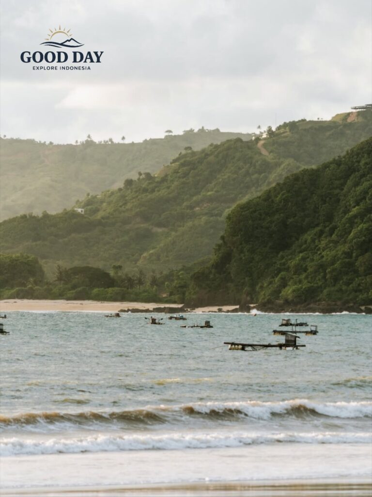 Lombok beach with gentle waves, traditional fishing platforms floating offshore, and lush green hills rising along the coastline under a soft tropical sky in West Nusa Tenggara, Indonesia.