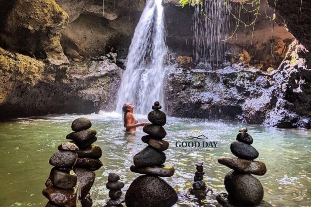 Hidden waterfall in Lombok surrounded by natural rock formations and a tranquil emerald pool, where a traveler enjoys a refreshing swim beneath cascading water. Stone cairns in the foreground add to the peaceful atmosphere of this secluded tropical paradise.
