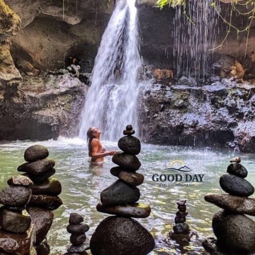 Hidden waterfall in Lombok surrounded by natural rock formations and a tranquil emerald pool, where a traveler enjoys a refreshing swim beneath cascading water. Stone cairns in the foreground add to the peaceful atmosphere of this secluded tropical paradise.