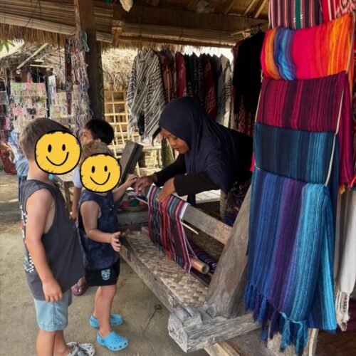 Children watching a local Sasak artisan demonstrate traditional hand weaving in Sade Traditional Village, Lombok, Indonesia, surrounded by colorful handmade textiles and authentic village craftsmanship.