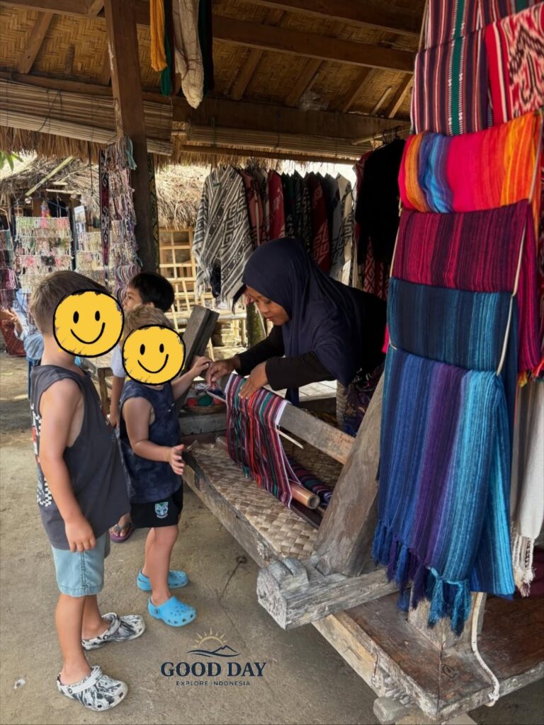 Children watching a local Sasak artisan demonstrate traditional hand weaving in Sade Traditional Village, Lombok, Indonesia, surrounded by colorful handmade textiles and authentic village craftsmanship.