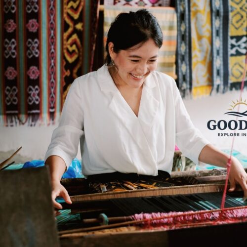 Visitor learning traditional Sasak hand-weaving techniques at Sade Traditional Village in Lombok, Indonesia, using a wooden loom surrounded by colorful handwoven textiles and authentic local craftsmanship.
