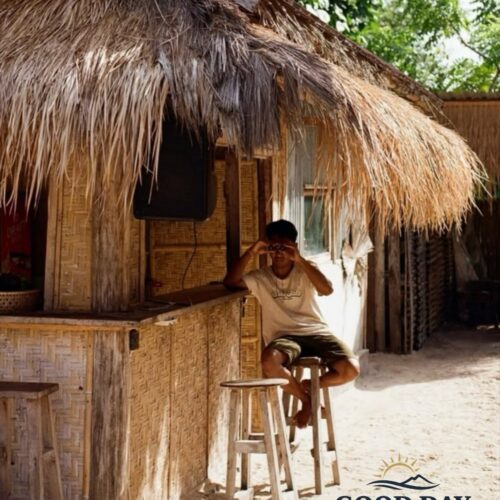 Traditional beachside bamboo hut at Tanjung Aan Beach, Lombok, Indonesia, with a local resident relaxing under a thatched roof surrounded by white sand and tropical island atmosphere.