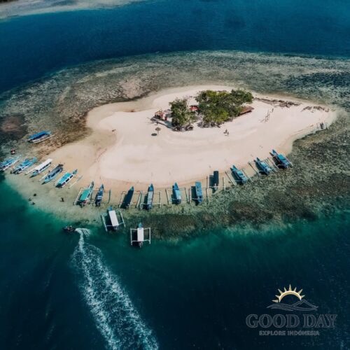 Aerial drone view of a small tropical Gili island in Lombok surrounded by turquoise waters, white sandy beaches, coral reefs, and traditional Indonesian outrigger boats anchored along the shoreline.