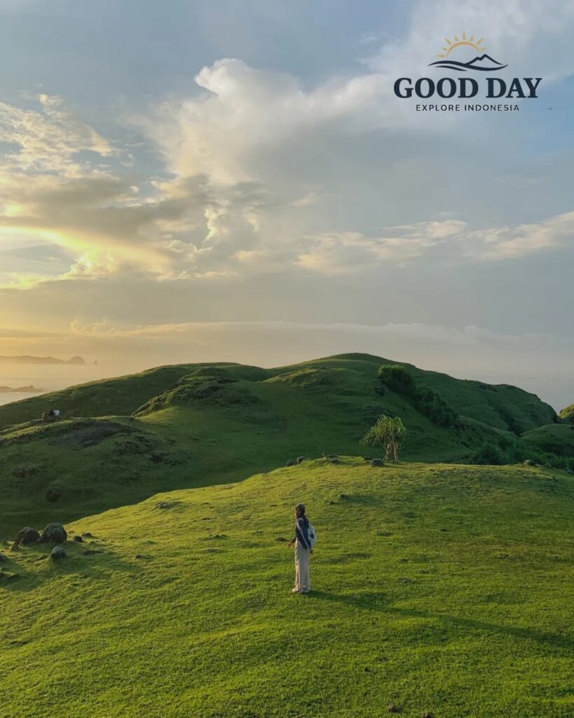 Traveler standing on the rolling green hills of Merese Hill in Lombok, Indonesia, during golden hour. The scenic landscape features lush grass-covered hills, dramatic clouds, soft sunset light, and panoramic views of the coastline near Kuta Mandalika.