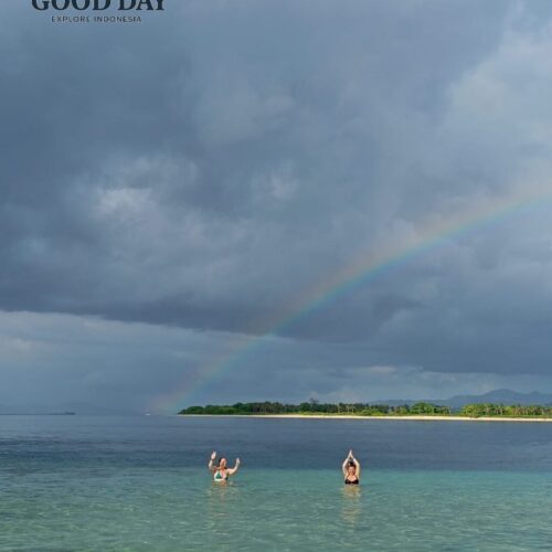 Two travelers enjoying the crystal-clear waters of Gili Nanggu, Lombok, beneath a dramatic sky and a beautiful rainbow stretching across the horizon. The pristine beach and turquoise sea create a breathtaking tropical island paradise.