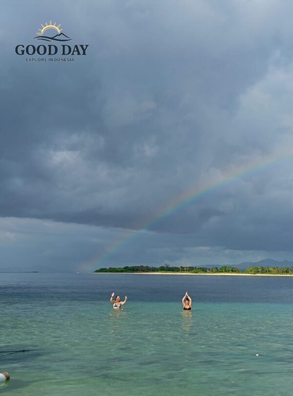 Two travelers enjoying the crystal-clear waters of Gili Nanggu, Lombok, beneath a dramatic sky and a beautiful rainbow stretching across the horizon. The pristine beach and turquoise sea create a breathtaking tropical island paradise.