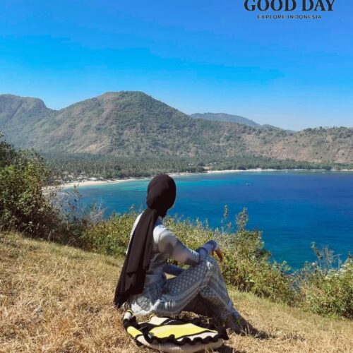 Traveler sitting on a grassy hilltop at Malibu Hill Lombok, Indonesia, overlooking a stunning turquoise bay, tropical coastline, and lush mountains under a clear blue sky. This scenic viewpoint offers one of the most breathtaking panoramic ocean views in Lombok.