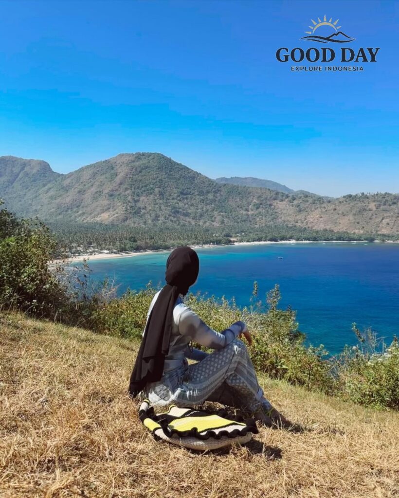 Traveler sitting on a grassy hilltop at Malibu Hill Lombok, Indonesia, overlooking a stunning turquoise bay, tropical coastline, and lush mountains under a clear blue sky. This scenic viewpoint offers one of the most breathtaking panoramic ocean views in Lombok.