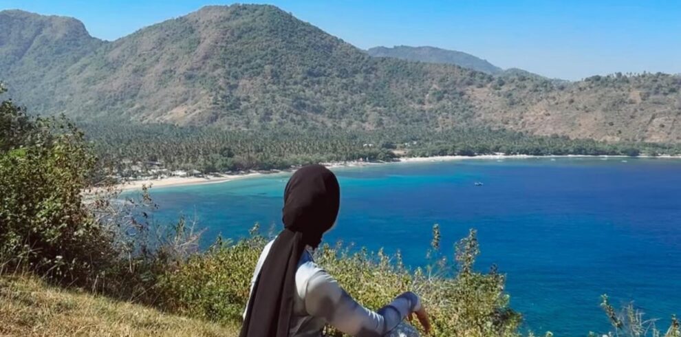 Traveler sitting on a grassy hilltop at Malibu Hill Lombok, Indonesia, overlooking a stunning turquoise bay, tropical coastline, and lush mountains under a clear blue sky. This scenic viewpoint offers one of the most breathtaking panoramic ocean views in Lombok.